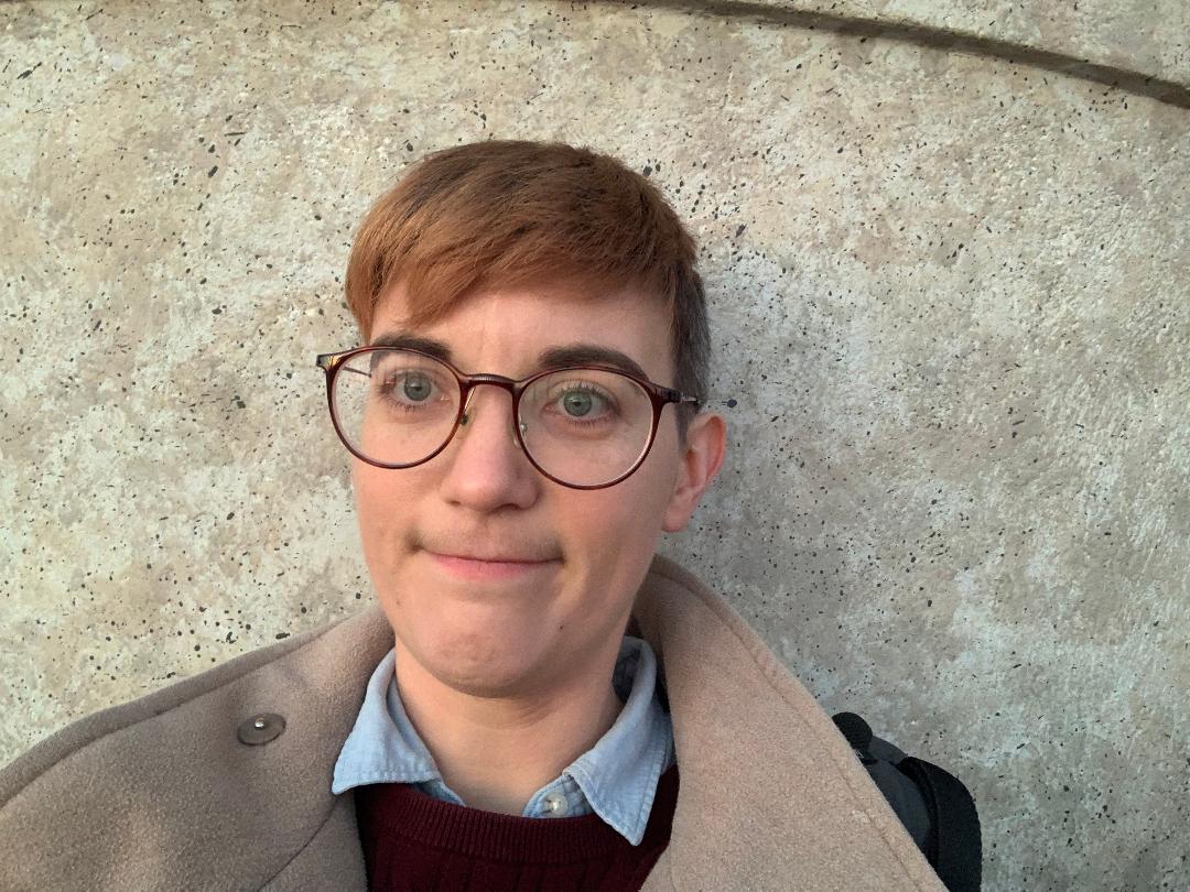 A photo of me: mid-30s nonbinary person with short reddish-brown hair, glasses, blue shirt and burgundy jumper against a stone wall background